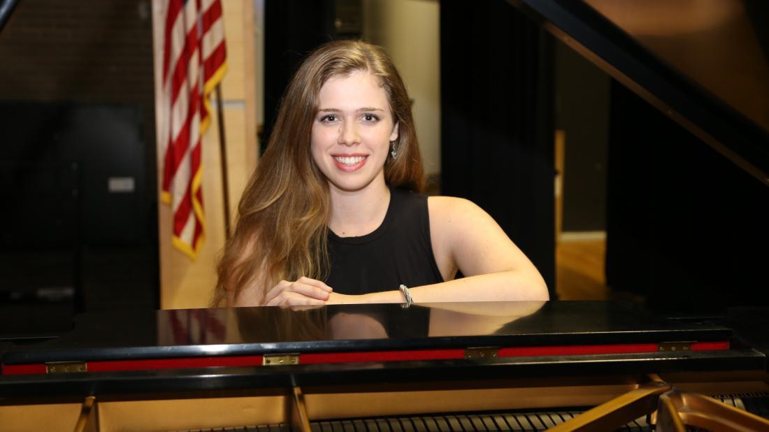 Woman in black dress poses at black baby grand piano on stage with American flag in background