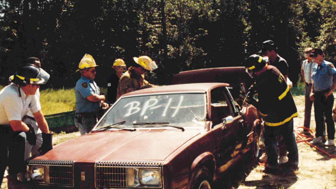 Male students working on decommissioned car outside during fire & rescue training in 1990