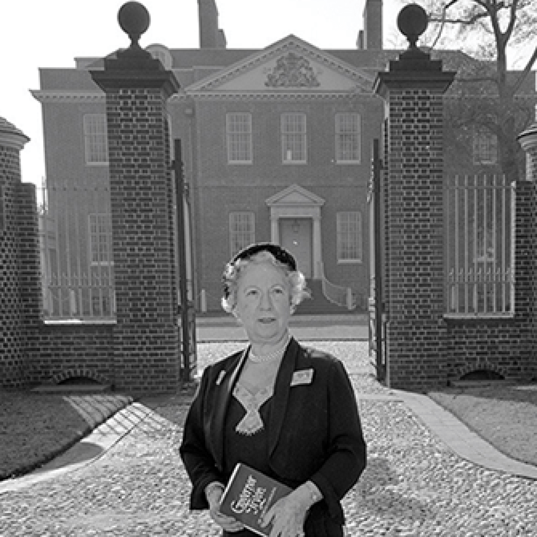 Older woman holding a book and standing near the front gate of Tryon Palace. Caption reads: Gertrude Carraway, journalist, historian, director, teacher, speaker, organizer, mover, and shaker as well as a leader in the effort to rebuild Tryon Palace.