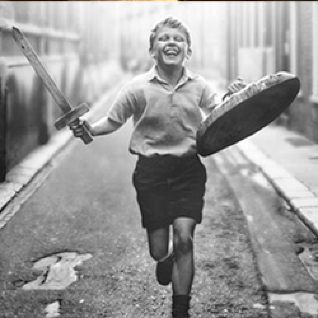 Black-and-white still of little boy running with a toy wooden sword and trash-can lid shield from the film Belfast