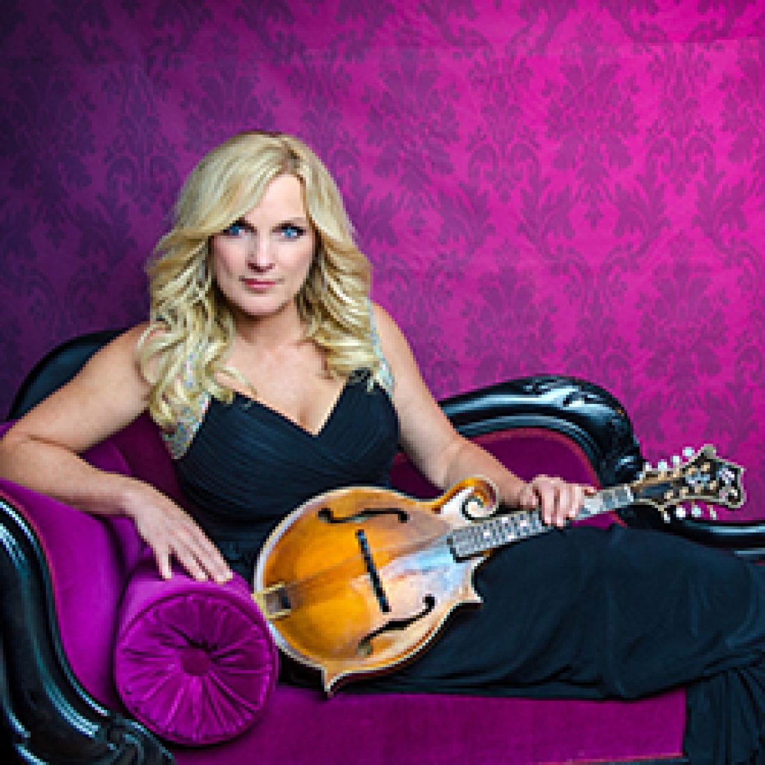 Country singer Rhonda Vincent poses holding a mandolin on a fuchsia couch in front of patterned fuchsia wallpaper