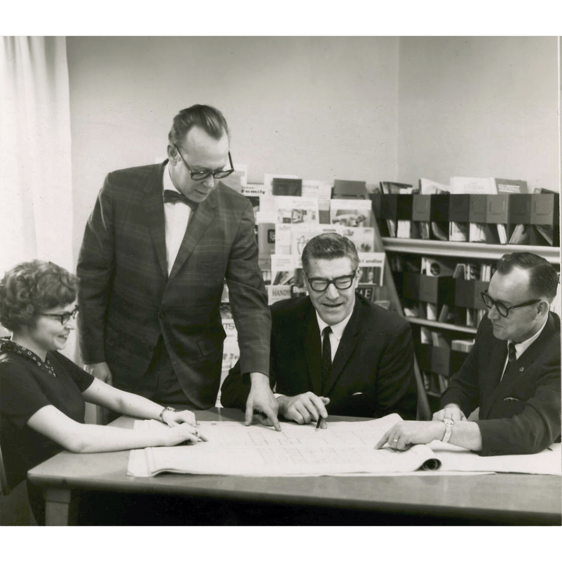 Black and white photo of three men and one woman sitting at a table looking at building plans