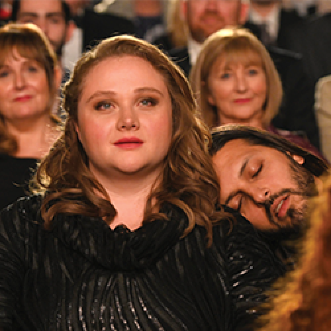 Audience of fancily dressed opera-goers are seated at a performance, with a man sleeping on a woman's shoulder