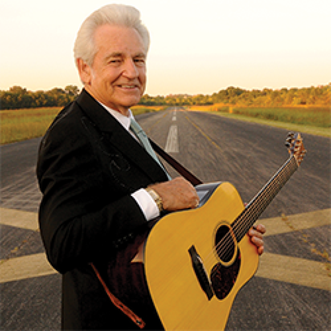 Older man wearing an acoustic guitar standing on a road in the country