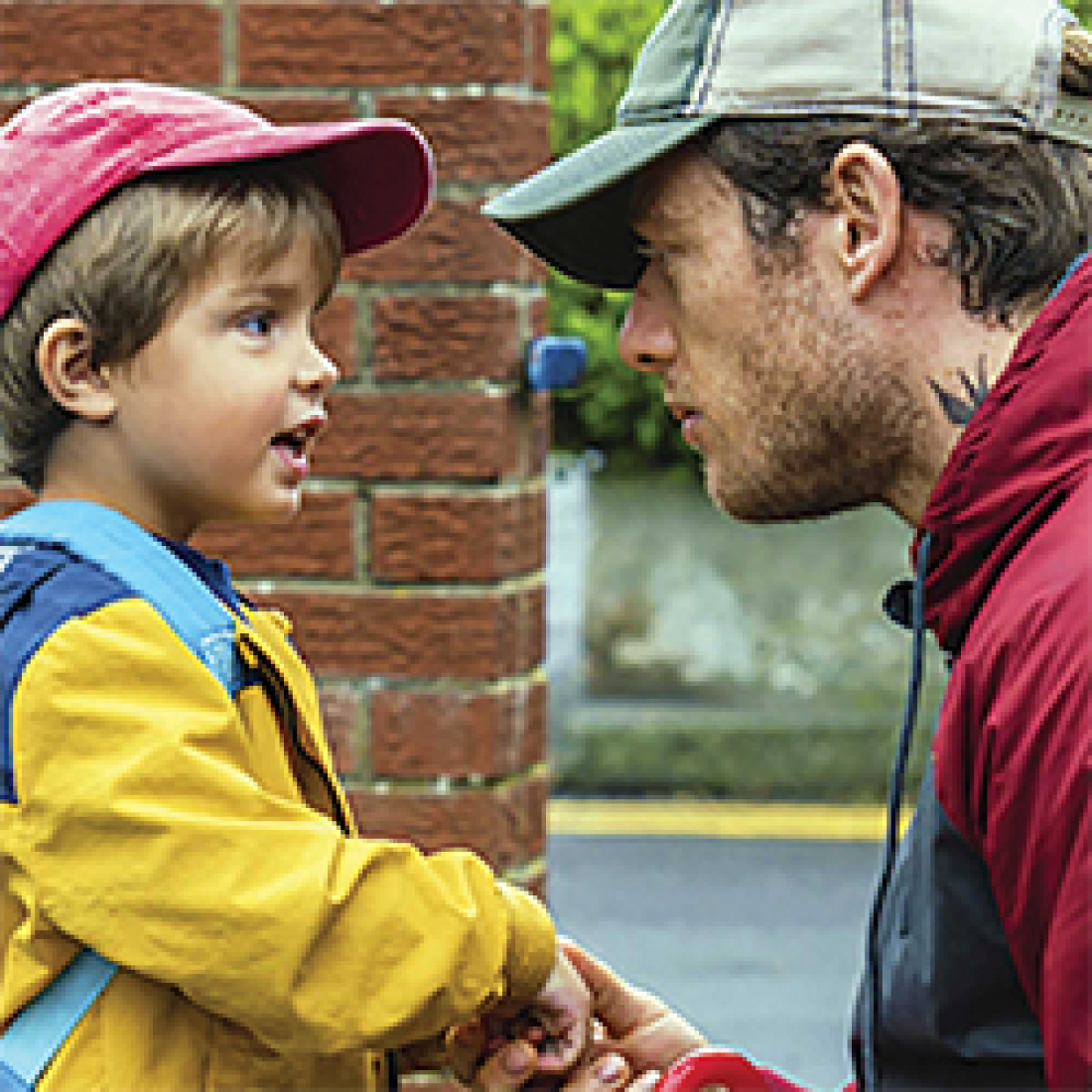 A young boy in red cap, yellow raincoat, and backpack talks to a men in a gray cap and red hoodie