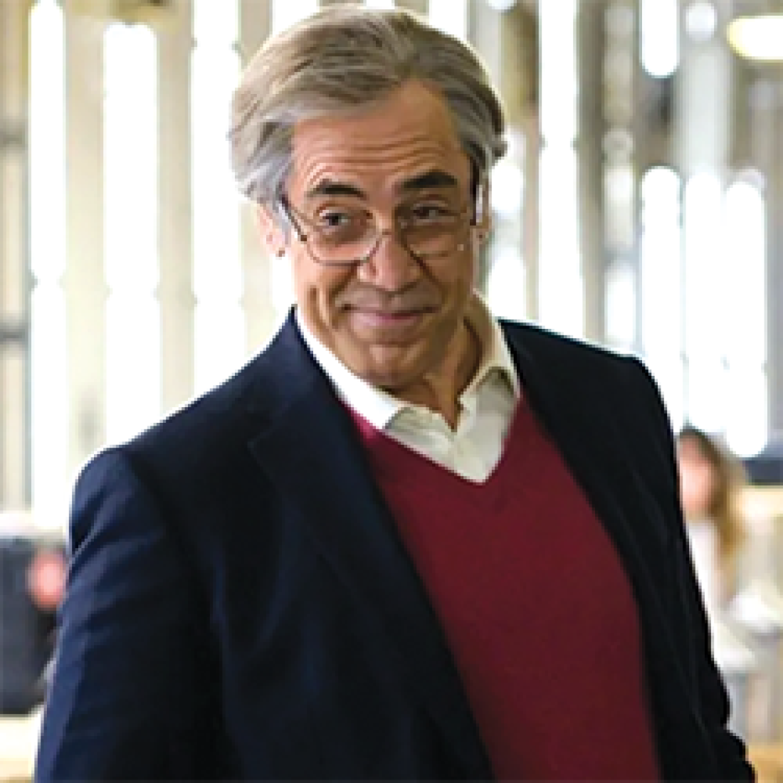 Older gentleman in glasses, white collared shirt, red sweater vest, and navy blue jacket smirks