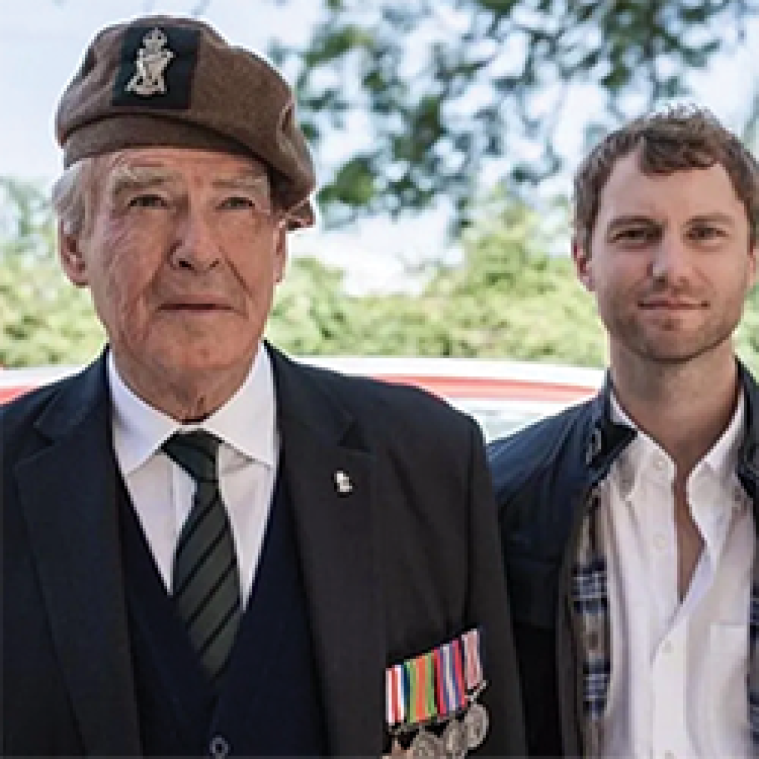 Older WWII veteran in uniform stands next to younger male outside