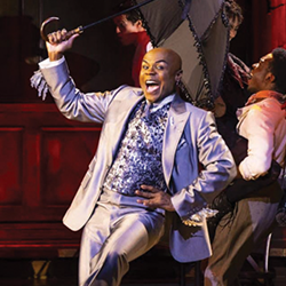Performer in a silver suit sings and dances with an umbrella during a lively scene from Hadestown.