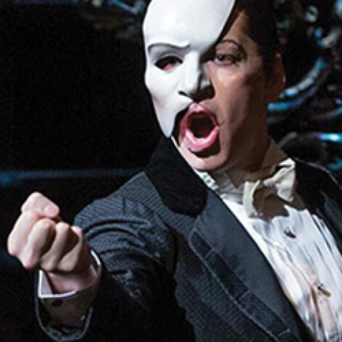 The Phantom, wearing his iconic white half-mask and black formal attire, gestures dramatically during a scene from The Phantom of the Opera.