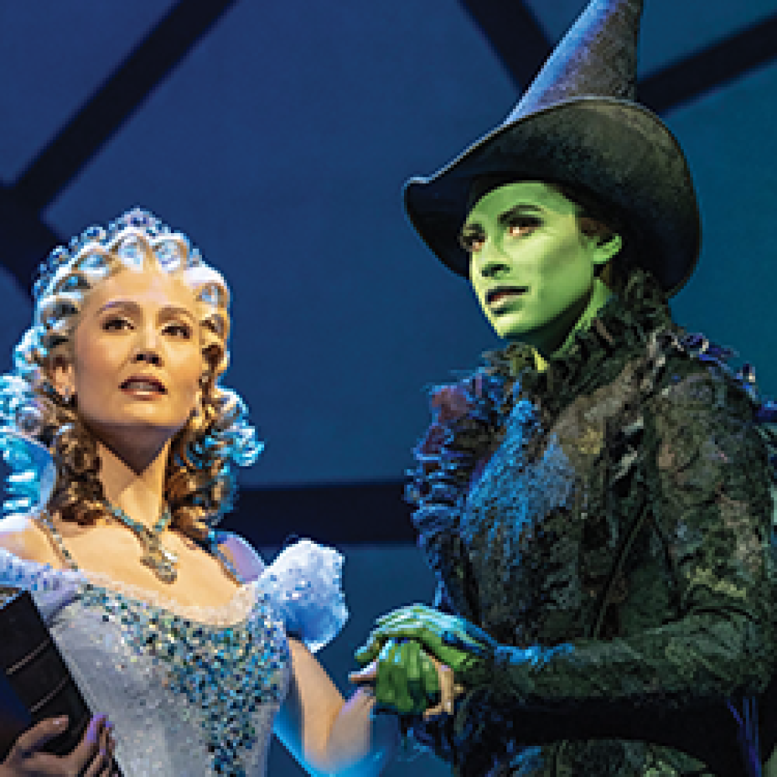 Glinda, dressed in a sparkling light-blue gown, stands beside the green-skinned Elphaba in a black dress and pointed hat during a scene from Wicked.