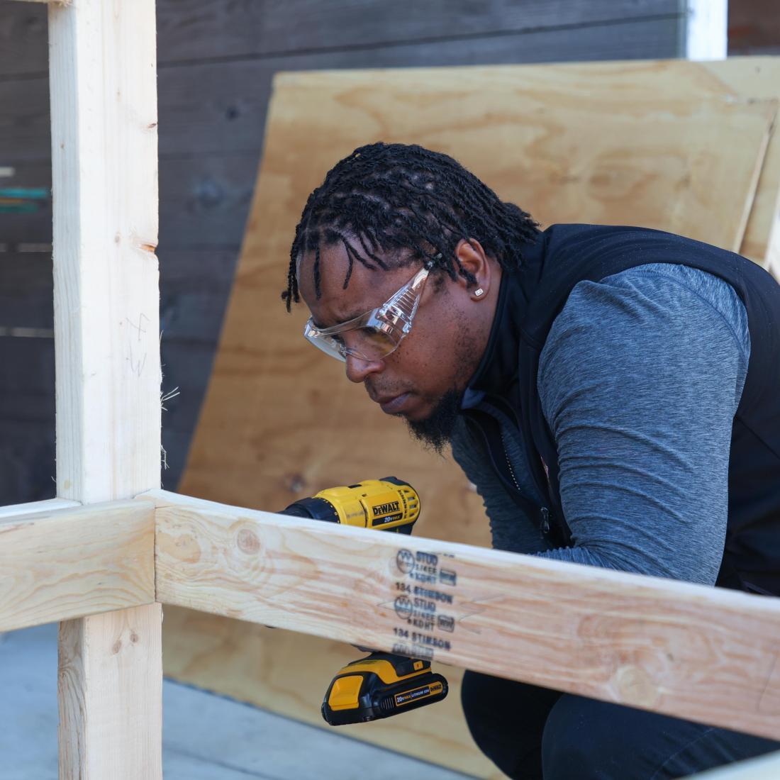 Construction student wearing safety glasses uses a drill to assemble a wooden frame.