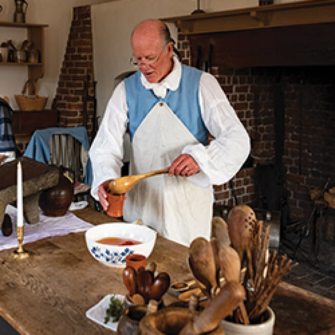 Historical interpreter in colonial clothing demonstrates food preparation at a wooden table in front of an open hearth.