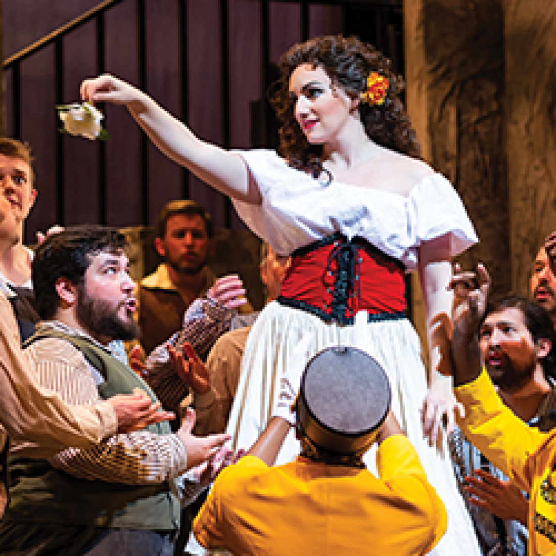 Carmen, dressed in a white blouse and red-and-black corset, dances above a crowd of men during a lively scene from Bizet’s Carmen.