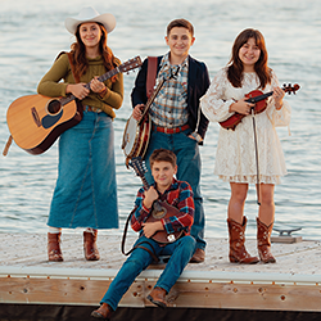 Four musicians pose by the water with acoustic instruments, including guitar, banjo, fiddle, and mandolin.