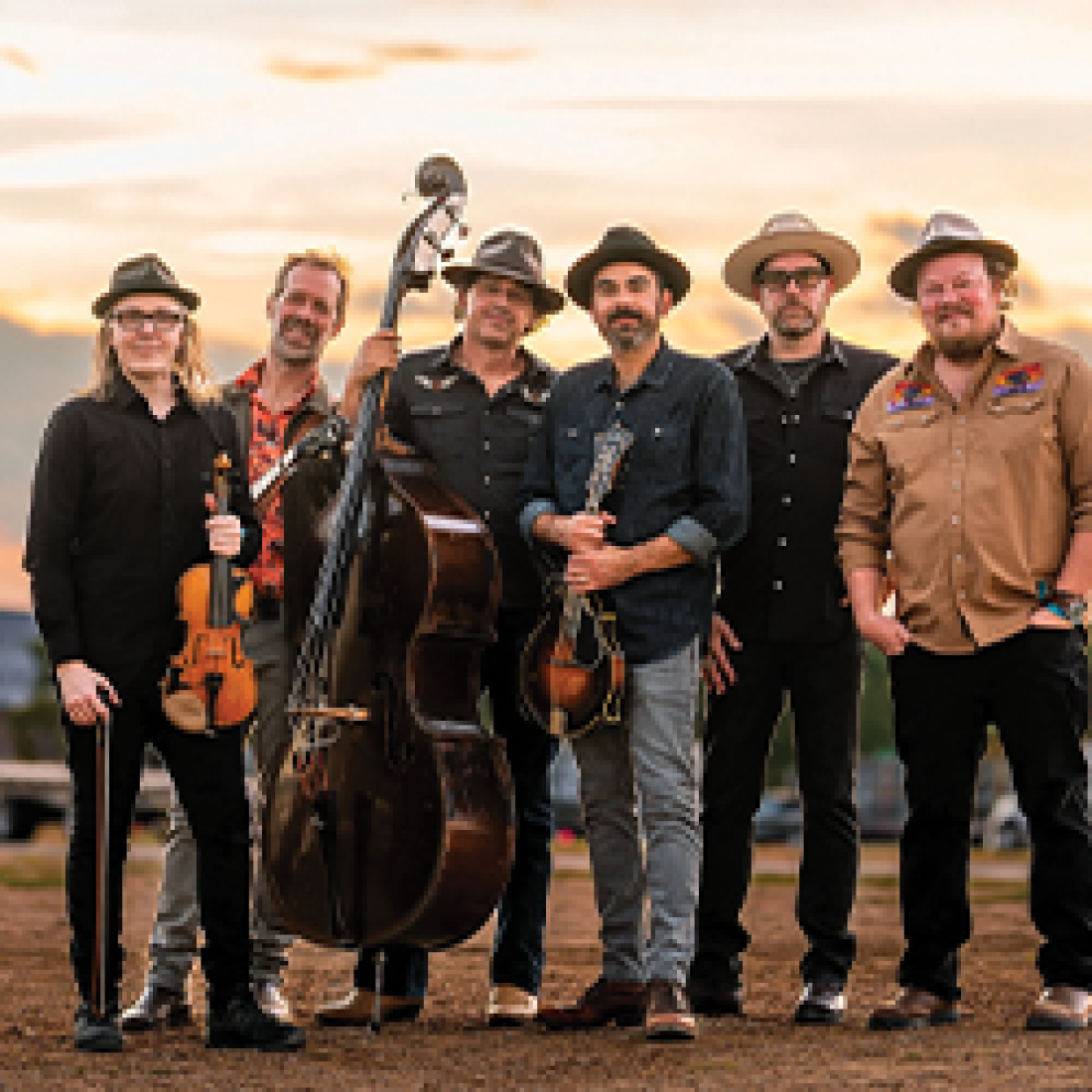 Seven musicians pose outdoors with bluegrass instruments, including fiddles, an upright bass, and a mandolin.