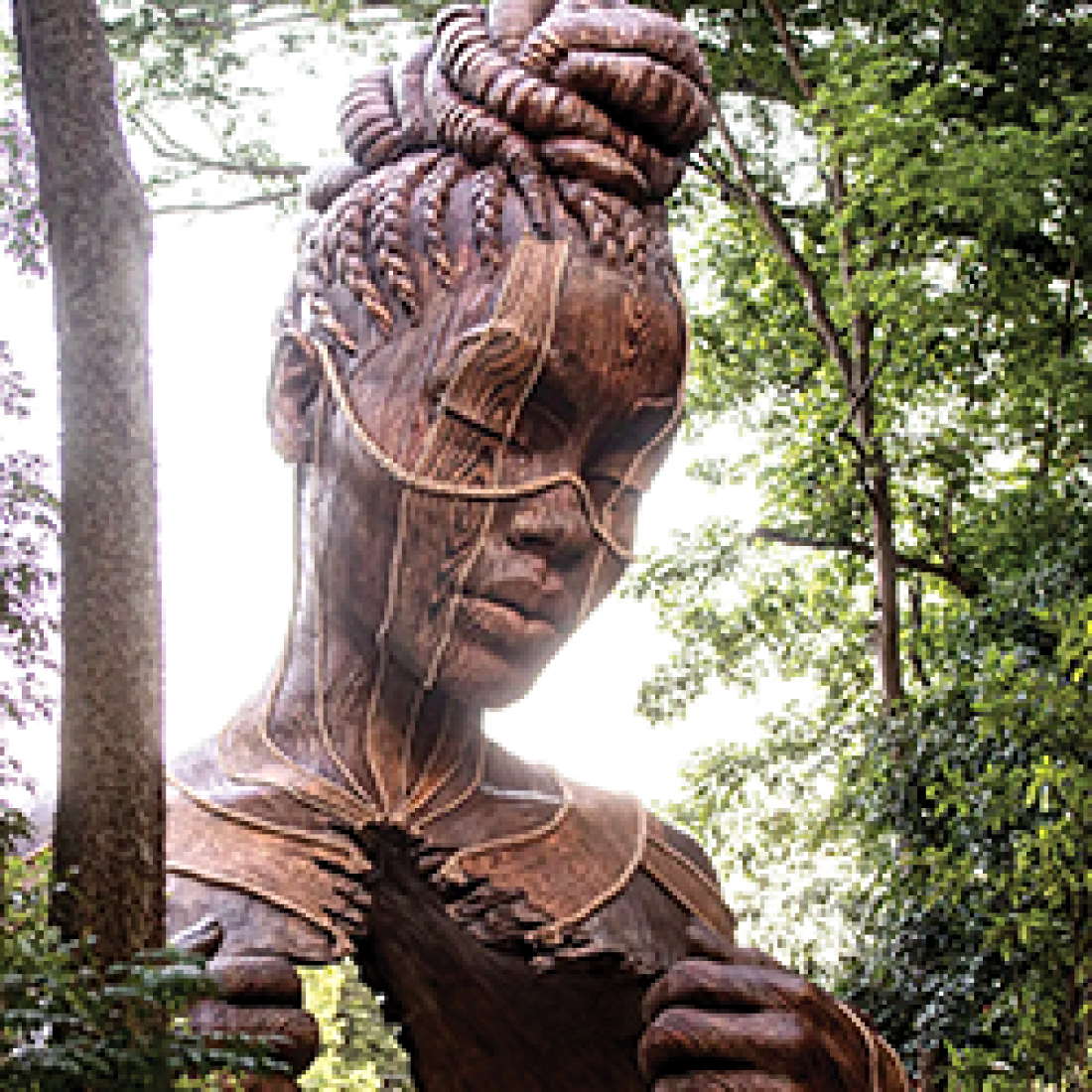 Large wooden sculpture of a woman with intricately braided hair stands among trees and appears to pull open a section of her chest.