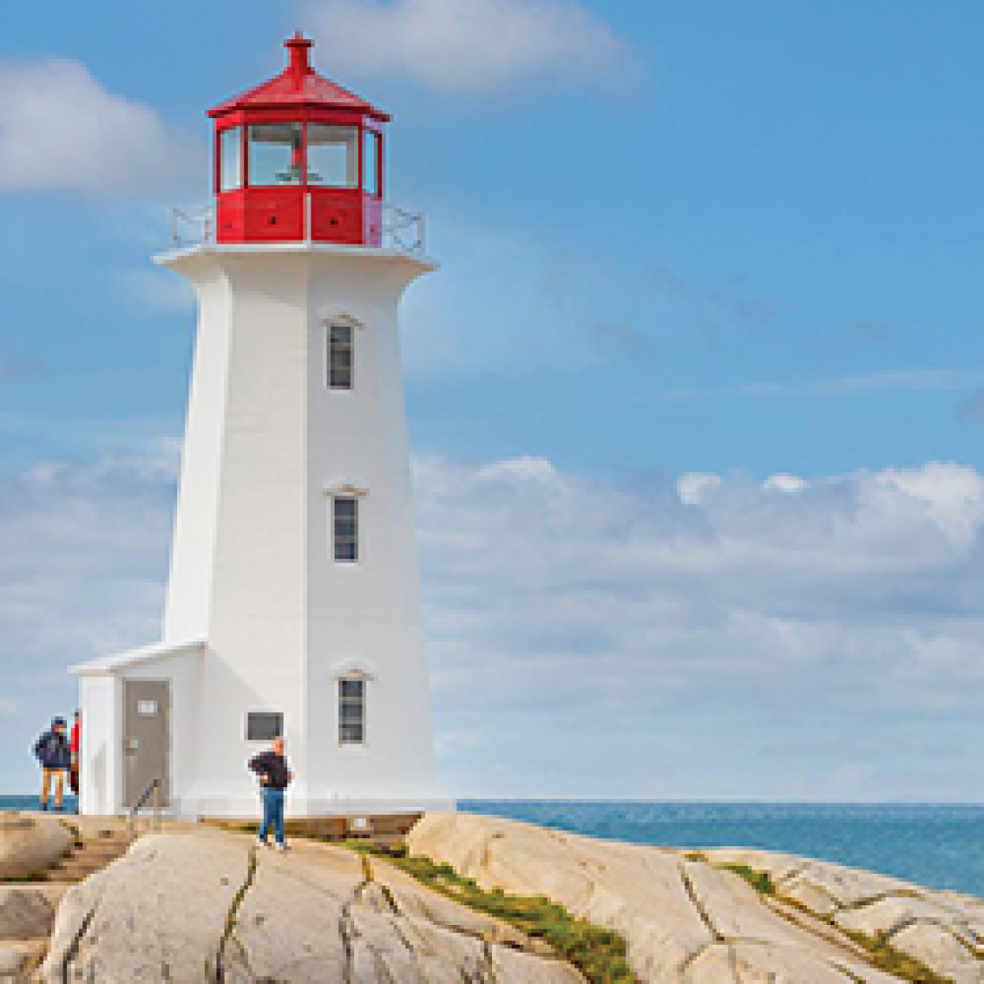 White lighthouse with a red lantern room stands on a rocky coastline overlooking the ocean in Nova Scotia.