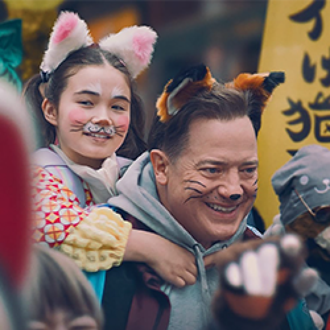 Smiling man carries a young girl wearing cat ears and face paint on his back during a lively festival scene from Rental Family.