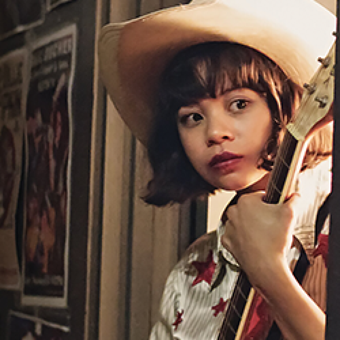 Young woman in a white cowboy hat holds a guitar and looks over her shoulder in a scene from Yellow Rose.