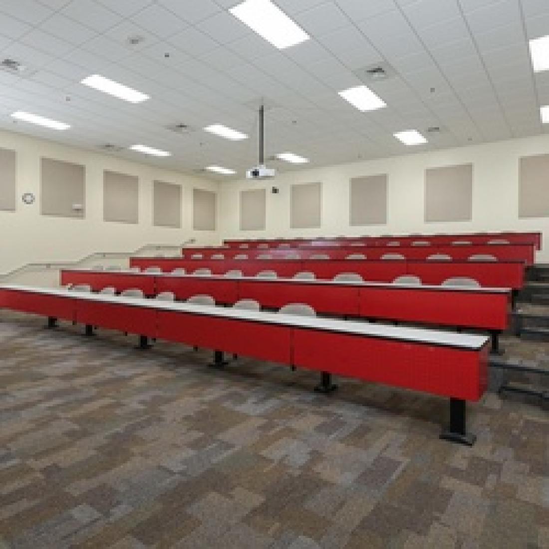 Tiered lecture hall with rows of red-fronted desks and gray chairs, a ceiling-mounted projector, side stairways, and a wooden podium at the front.