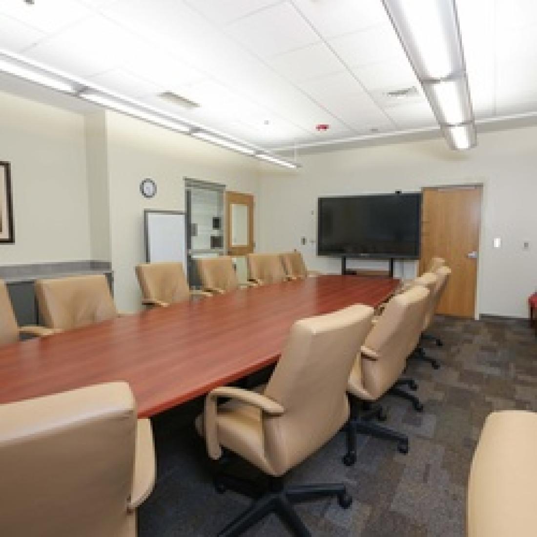 Conference room with a large wood table surrounded by tan chairs, additional red guest chairs along the wall, and a large interactive display at the front of the room.
