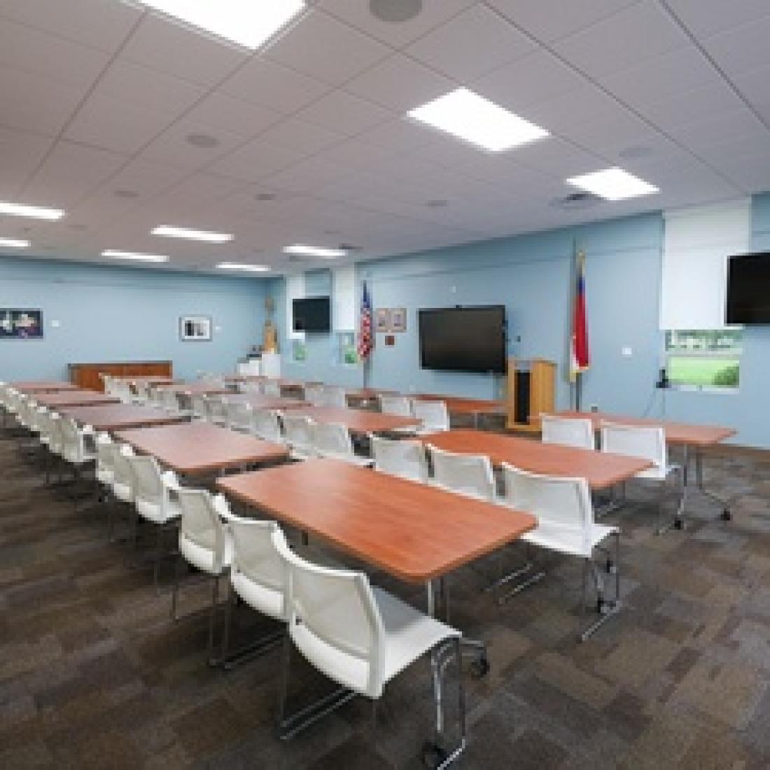 Bright meeting room with rows of tables and white chairs facing presentation screens and a podium, with blue walls, windows, and additional seating along the side.