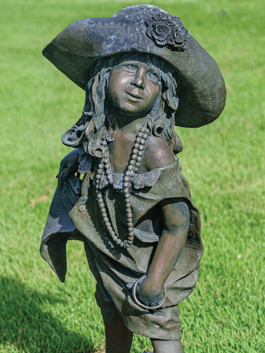 Bronze statue of little girl dressed in her mother's hat, shoes, and pearls