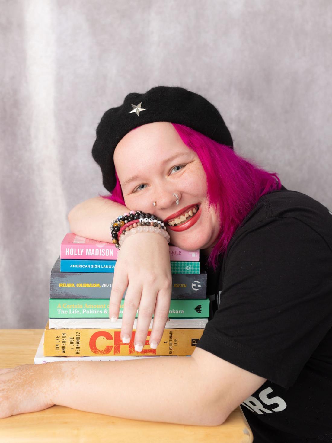 Craven alum Xena Casey smiles and leans over a pie of textbooks, wearing a black beret with silver star, black T-shirt, and bright pink hair