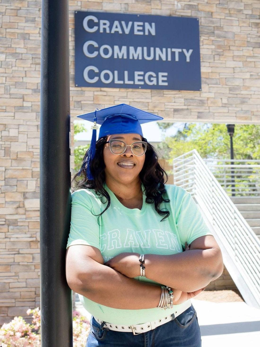 Craven alum Catrina Mack Cook crosses her arms and wears a grad cap in front of the Craven Community College sign