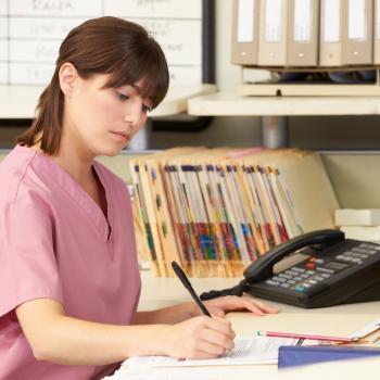Medical Office Administration worker writes notes at a desk with files and computer and phone