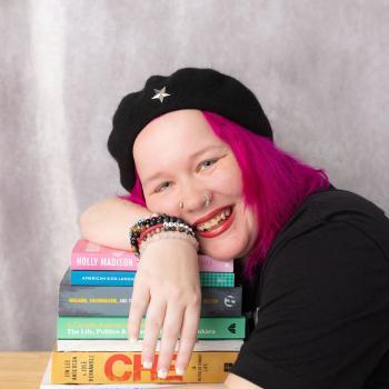 Craven alum Xena Casey smiles and leans over a pie of textbooks, wearing a black beret with silver star, black T-shirt, and bright pink hair