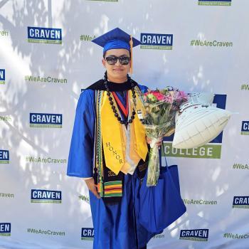 Craven CC alum Dominic Bradfield poses against Craven CC backdrop in cap and gown holding a bouquet of flowers and a balloon