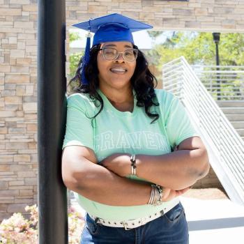 Craven alum Catrina Mack Cook crosses her arms and wears a grad cap in front of the Craven Community College sign