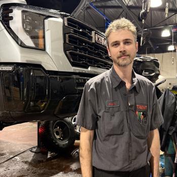 Automotive Systems Technology graduate Adam Heckman stands in front of a suspended car in the shop
