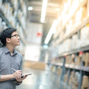 Young male worker taking stock of products in cardboard boxes on shelves in warehouse using digital tablet and pen