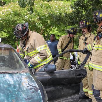 Firefighting students in protective gear practice an emergency response around a vehicle.