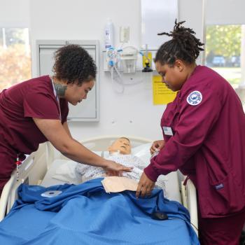 Nursing students in scrubs practice patient care using a medical training mannequin.