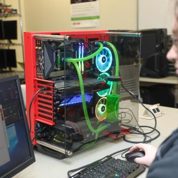 Information technology student works at a computer beside a custom-built PC with visible internal components and liquid cooling.