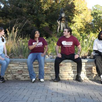 Four students sit spaced apart on a low stone wall in a landscaped campus courtyard, talking and relaxing outdoors.