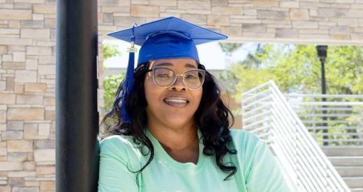 Craven alum Catrina Mack Cook crosses her arms and wears a grad cap in front of the Craven Community College sign