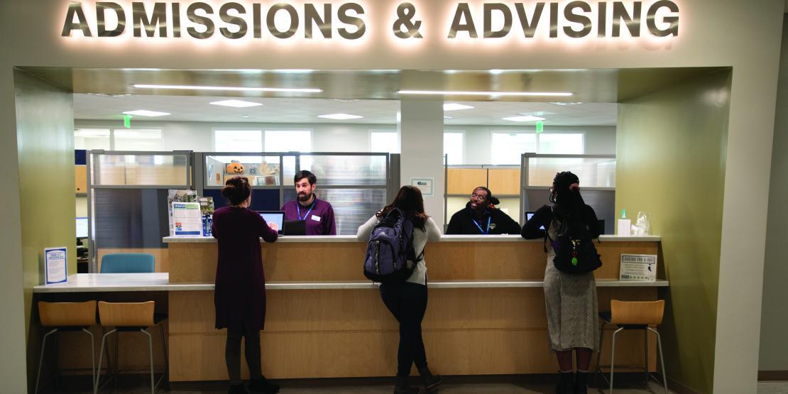 Three students stand at the counter while being helped by academic advisors under an illuminated sign reading "Admissions & Advising"