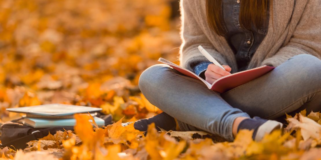 Student sitting criss-cross applesauce in orange fall leaves writing in notebook