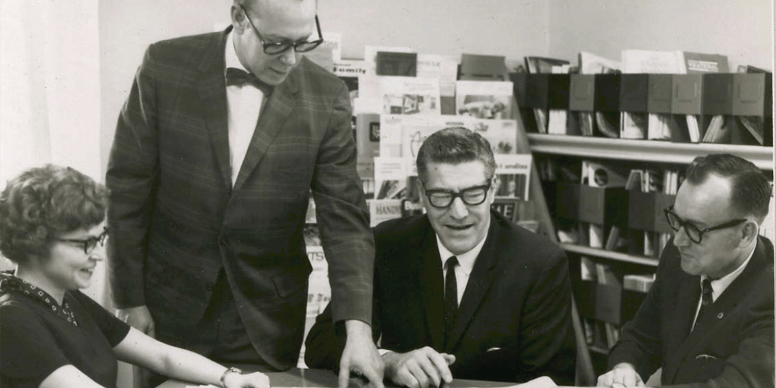 Black and white photo of three men and one woman sitting at a table looking at building plans