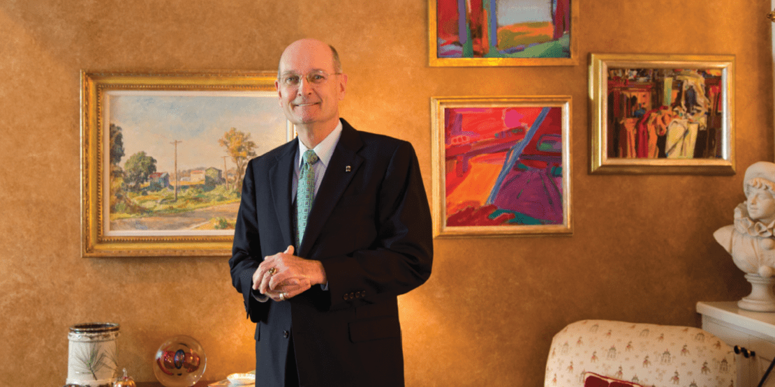 Dr. Jim Congleton wearing a suit and standing with hands clasped in a fancy foyer with paintings on the wall