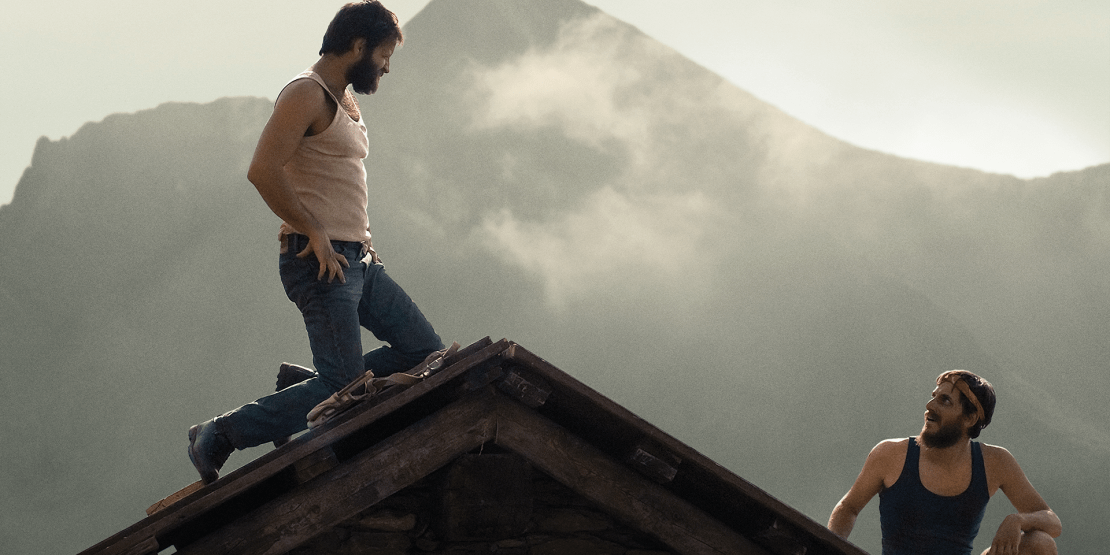 A man in jeans and white undershirt kneels on the apex of a hill with hands on hips, looking down at another man a few feet below on the other side of the hill