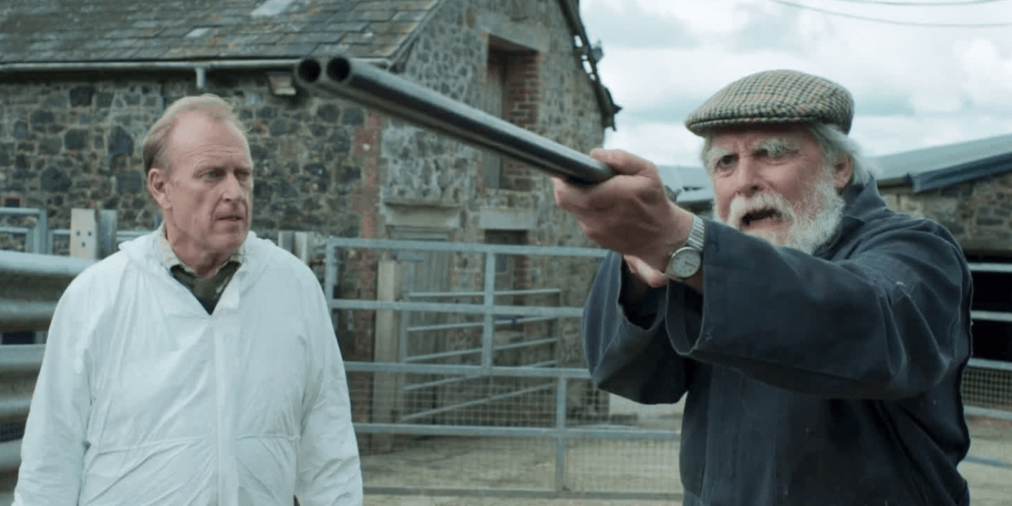 Man in white button-down shirt looking at man aiming rifle with old barn behind them