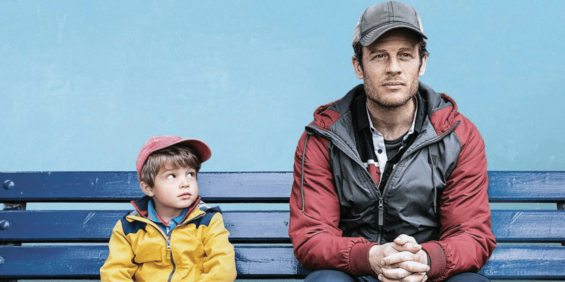 Young boy in red cap and yellow jacket looks over at middle-aged man with clasped hands as they both sit on a bench
