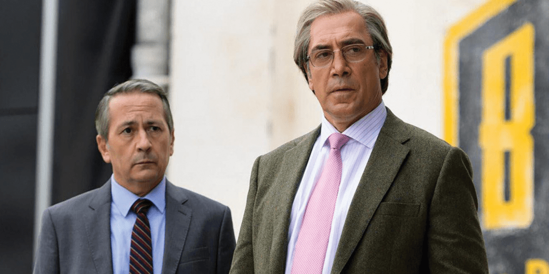 Older gentleman in glasses, light blue collared shirt, light pink tie, and sports coat and another older man in similar attire look ahead with serious expressions