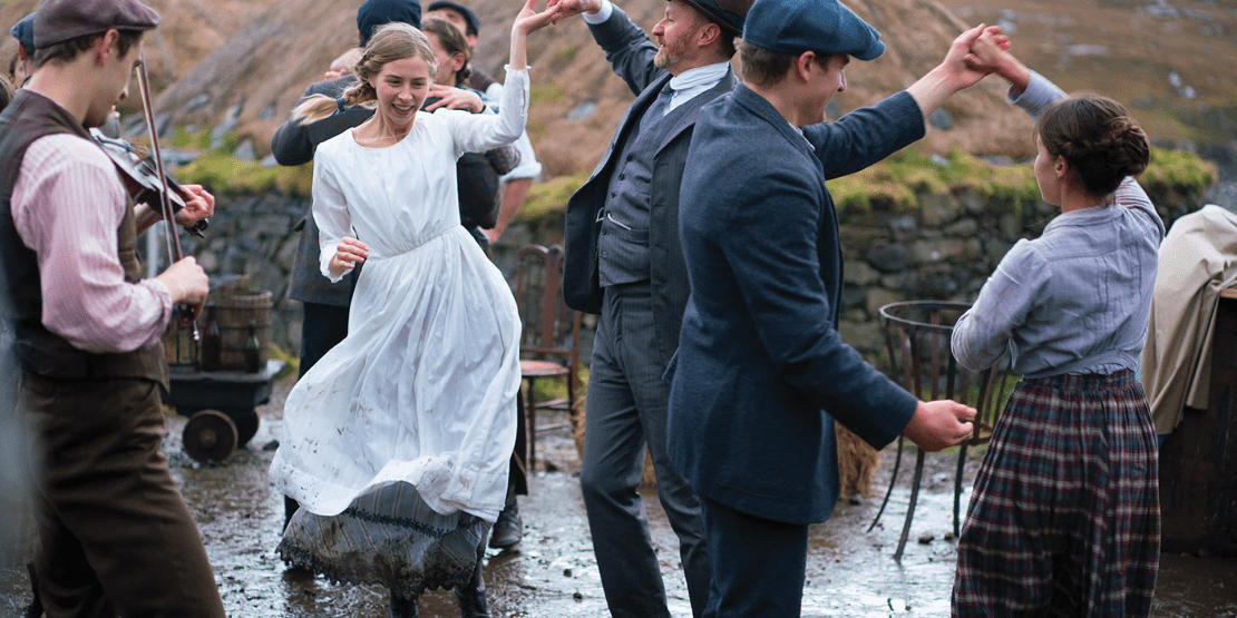 Woman in white dress dances in a shallow river with man, surrounded by other dancing couples