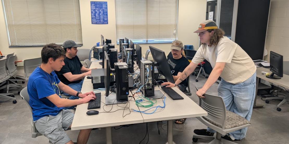 four students stand around a desk of computers